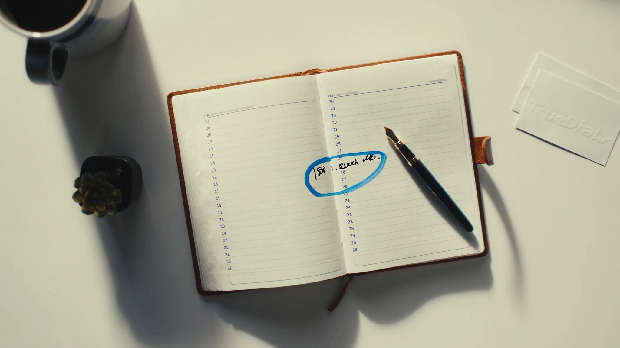 A leather-bound paper desk calendar open to a page with one time slot circled in brand-blue ink, a fountain pen resting on the page, a coffee mug, a small succulent